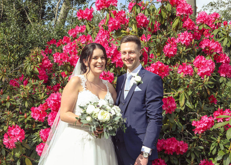 A bride and groom smiling stood in front of a pink azaela in full bloom on their wedding day at Haughley Park Barn Wedding Venue  photographed by Suffolk Wedding Photographers Hayley Denston Photography