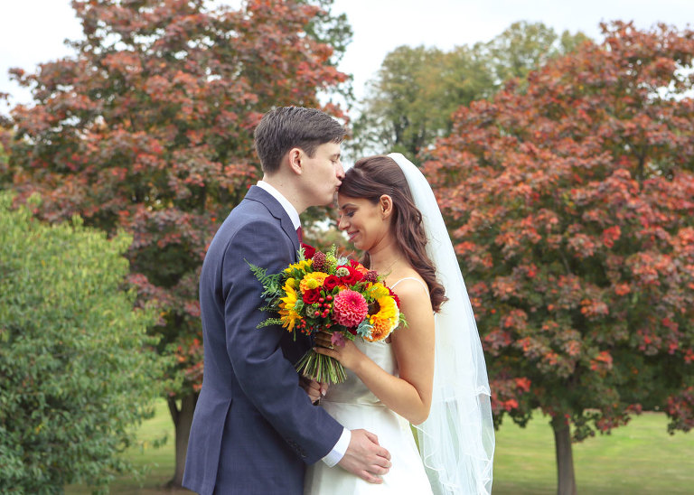 A groom kissing his bride on the head with dark red autumnal trees behind them and the brides bright autumnal wedding bouquet on their wedding day at Haughley Park Barn Wedding Venue photographed by Suffolk Wedding Photographers Hayley Denston Photography