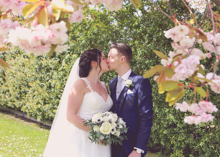 A bride and groom kissing underneath pale pink cherry blossom on their wedding day at Haughley Park Barn Wedding Venue photographed by Suffolk Wedding Photographers Hayley Denston Photography