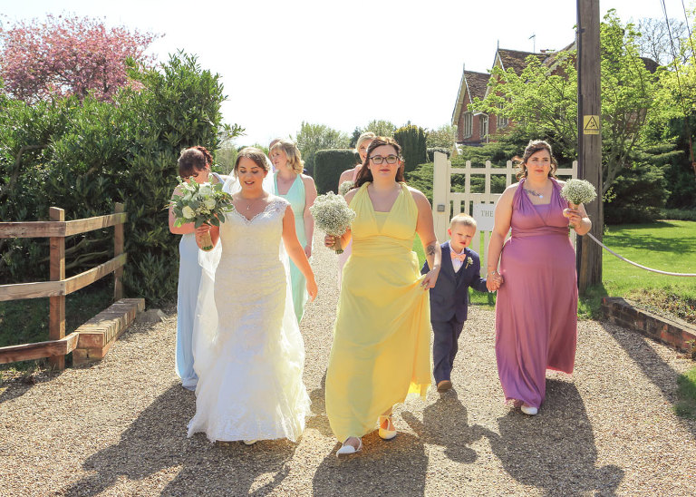 A bride and her bridesmaids and pageboy walking over to her wedding ceremony at Haughley Park Barn Wedding Venue photographed by Suffolk Wedding Photographers Hayley Denston Photography