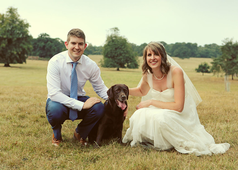 A bride and groom sat with their chocolate labrador in the parkland at Haughley Park Barn Wedding Venue on their wedding day photographed by Suffolk Wedding Photographers Hayley Denston Photography