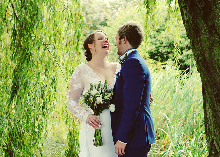 A bride and groom laughing underneath willow trees on their wet wedding day at Haughley Park Barn Wedding Venue photographed by Suffolk Wedding Photographers Hayley Denston Photography