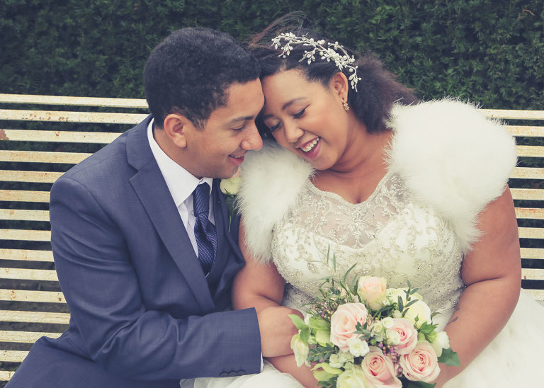 A bride and groom nuzzling together on their cool spring wedding day sat on a metal bench at Haughley Park Barn Wedding Venue on their wedding day photographed by Suffolk Wedding Photographers Hayley Denston Photography