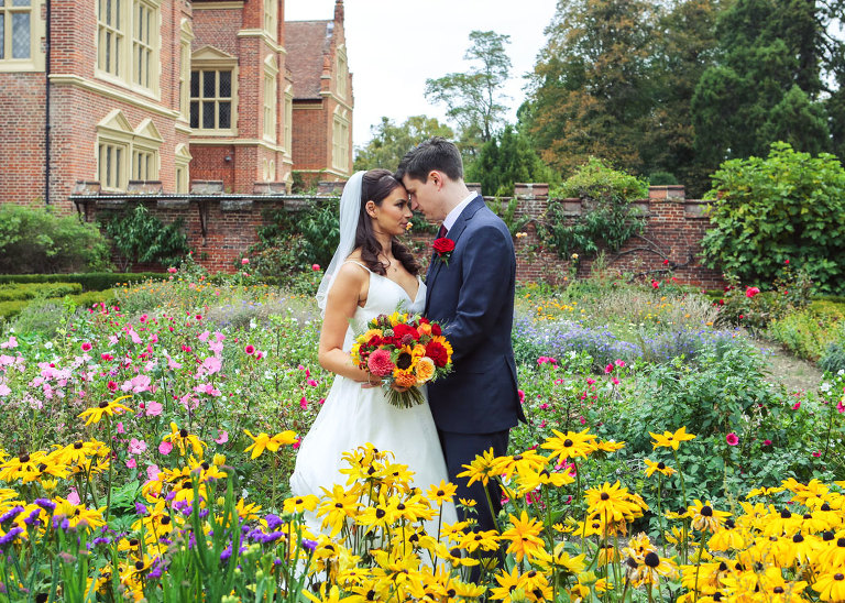 A bride and groom stood in bright flower beds for their autumnal wedding day at Haughley Park Barn Wedding Venue photographed by Suffolk Wedding Photographers Hayley Denston Photography