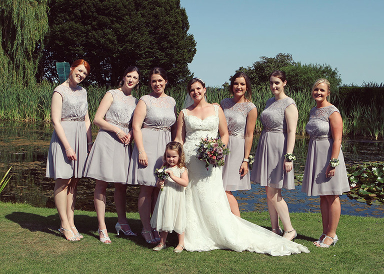 A bride and her bridesmaids and flowergirls stood in front of the pond at Haughley Park Barn Wedding Venue photographed by Suffolk Wedding Photographers Hayley Denston Photography