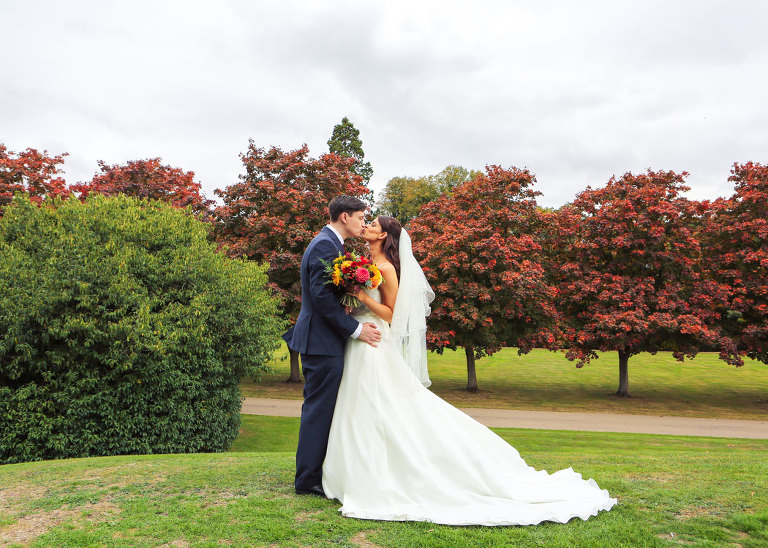 A bride and groom kissing on a hill with dark red autumnal trees behind them on their wedding day at Haughley Park Barn Wedding Venue photographed by Suffolk Wedding Photographers Hayley Denston Photography