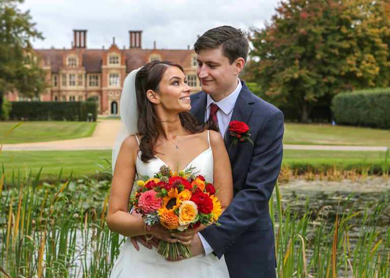 A bride and groom looking at each other with the pond and the hall behind them on their autumnal wedding day at Haughley Park Barn Wedding Venue photographed by Suffolk Wedding Photographers Hayley Denston Photography
