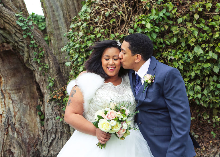 A groom kissing his bride in front of one of the oldest oak trees in Suffolk at Haughley Park Barn Wedding Venue photographed by Suffolk Wedding Photographers Hayley Denston Photography