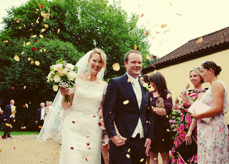 A bride and groom smiling and walking through a confetti aisle at Haughley Park Barn Wedding Venue photographed by Suffolk Wedding Photographers Hayley Denston Photography