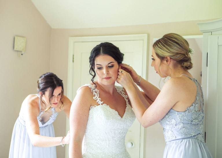 A bride having her earrings put in by a bridesmaid whilst another bridesmaid does her dress up at the Farmhouse at Haughley Park Barn Wedding Venue photographed by Suffolk Wedding Photographers Hayley Denston Photography