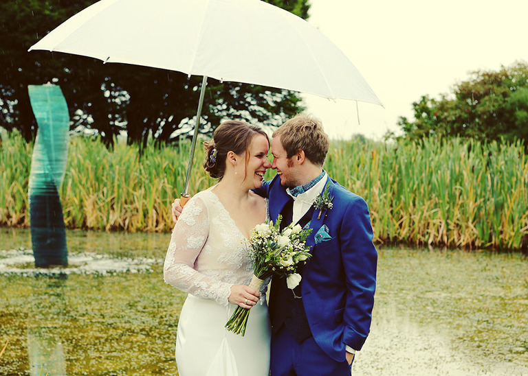 A bride and groom underneath a white umbrella laughing together on their wet wedding day at Haughley Park Barn Wedding Venue photographed by Suffolk Wedding Photographers Hayley Denston Photography