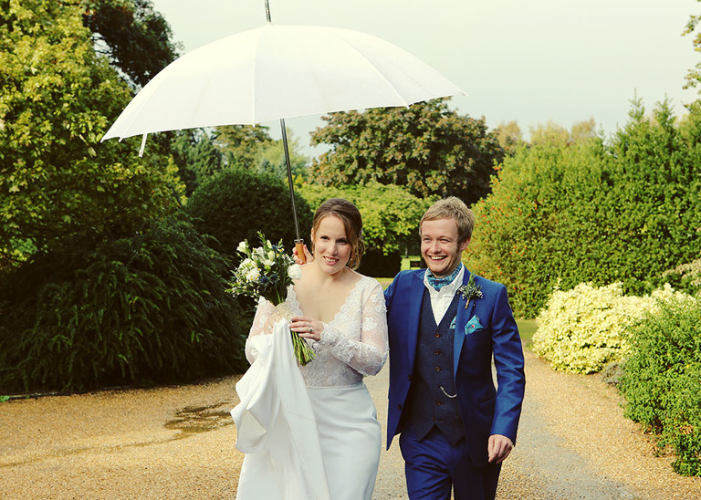 A bride and groom walking whilst holding a giant white umbrella on their wet wedding day at Haughley Park Barn Wedding Venue photographed by Suffolk Wedding Photographers Hayley Denston Photography