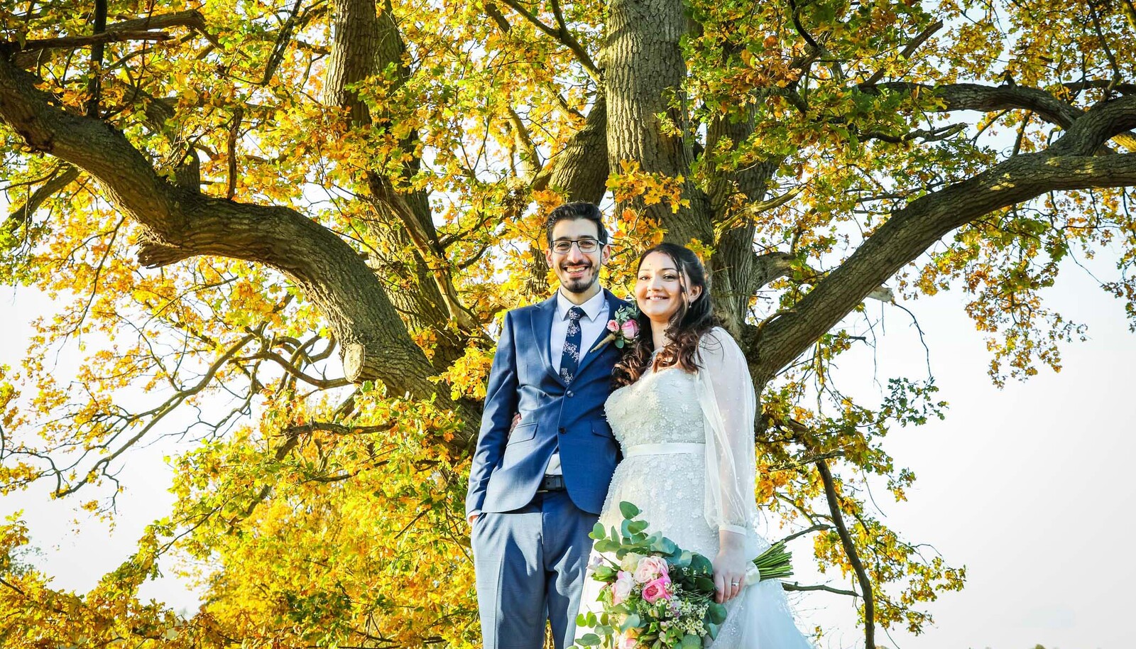 A bride and groom under a bright yellow autumnal oak tree on their wedding day at Easton Grange photographed by Suffolk Wedding Photographers Hayley Denston Photography