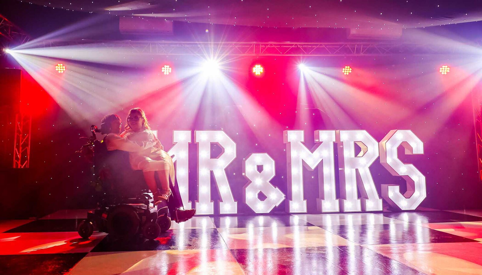 A bride and groom on their wedding day with the groom in his wheelchair during thier first dance with lots of disco lights and a big lit Mr and Mrs sign at All Manor of Events Wedding Venue photographed by Suffolk Wedding Photographers Hayley Denston Photography
