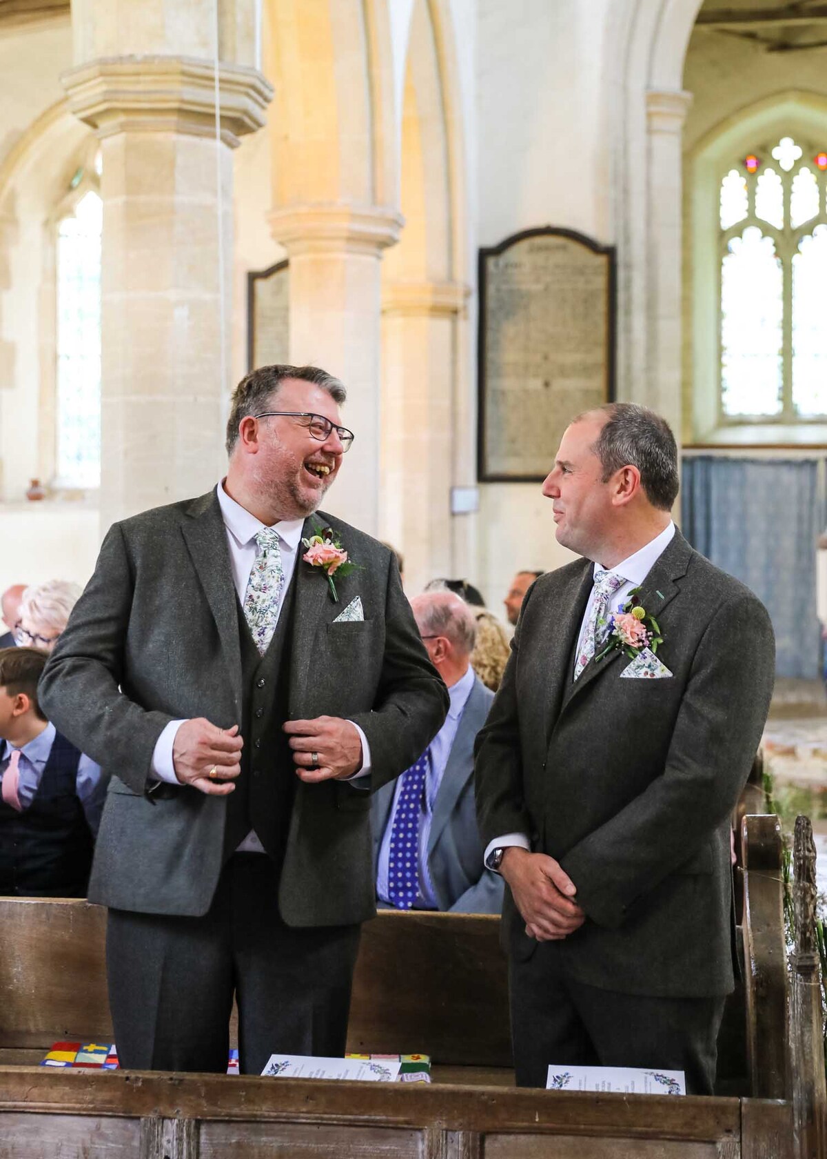 A groom and his bestman laughing at the front of a suffolk church before the wedding ceremony captured by Suffolk Wedding Photographers Hayley Denston Photography