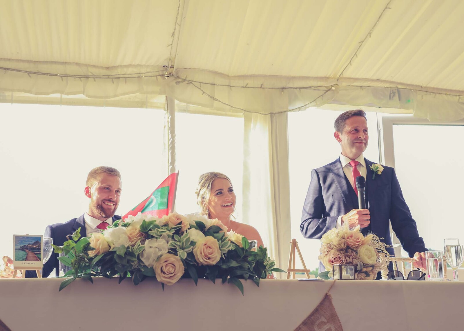 A father of the bride and the bride and groom during speeches in a marquee captured by Suffolk Wedding Photographers Hayley Denston Photography