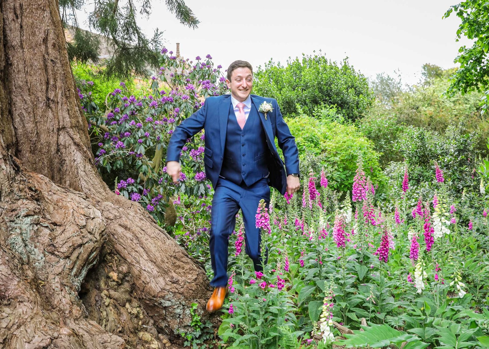 A groom jumping out of foxgloves in the wood at Haughley Park Barn Wedding Venue photographed by Suffolk Wedding Photographers Hayley Denston Photography