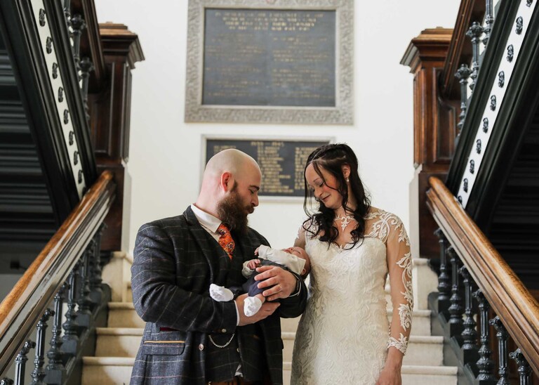 A bride and groom looking at their baby on the stairs at Ipswich Town Hall on their wedding day captured by Suffolk Wedding Photographers Hayley Denston Photography