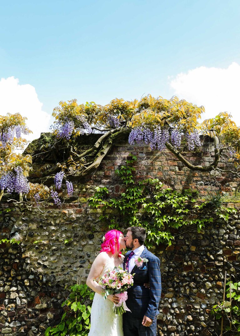 A bride and groom kissing in front of a flowering wisteria in the Abbey Gardens in Bury St Edmunds on a wedding day in Suffolk captured by Suffolk Wedding Photographers Hayley Denston Photography