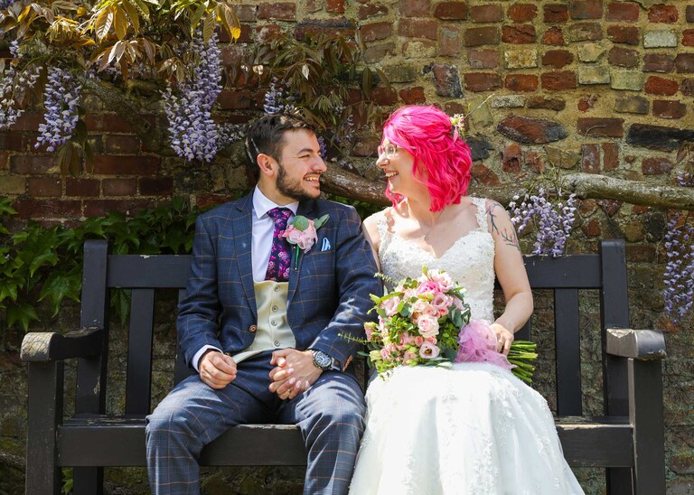 A bride and groom sat on a bench in front of wisteria in bloom in the Abbey Gardens in Bury St Edmunds on a wedding day in Suffolk captured by Suffolk Wedding Photographers Hayley Denston Photography