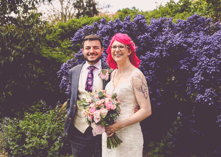 A bride and groom stood in front of a ceanothus bush in the Abbey Gardens in Bury St Edmunds on a wedding day in Suffolk captured by Suffolk Wedding Photographers Hayley Denston Photography