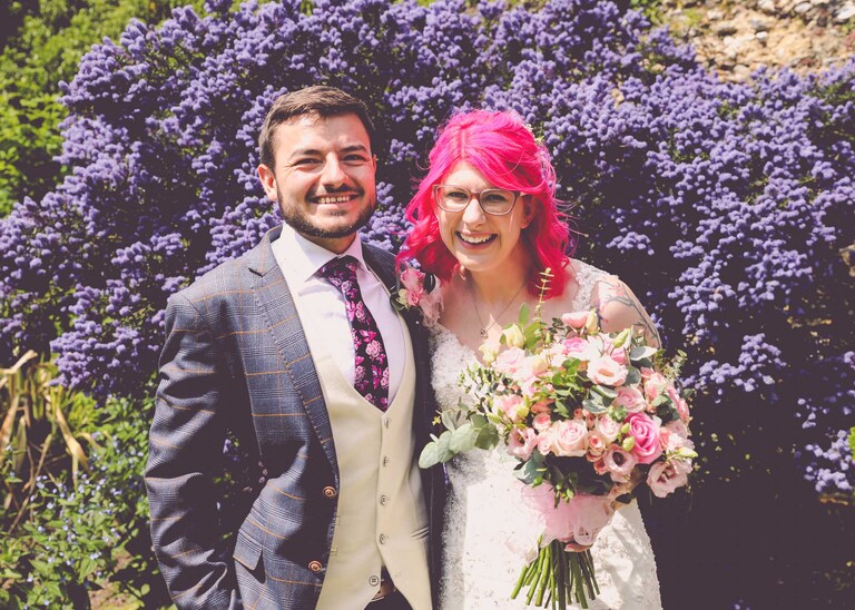 A bride and groom laughing in front of a blue ceanothus bush on their wedding day in the Abbey Gardens in Bury St Edmunds in Suffolk captured by Suffolk Wedding Photographers Hayley Denston Photography