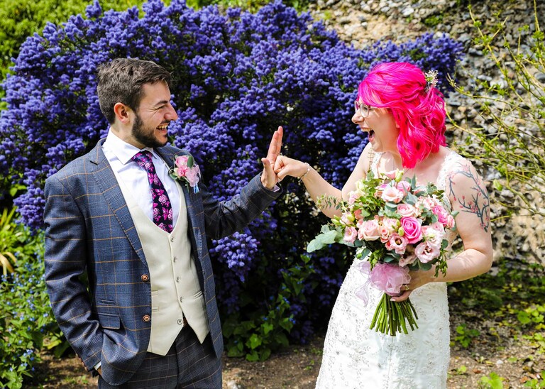 A bride and groom fist bumping in front of a bright blue ceanothus bush in the Abbey Gardens in Bury St Edmunds on a wedding day in Suffolk captured by Suffolk Wedding Photographers Hayley Denston Photography