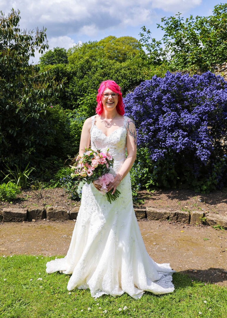 A bride stood in front of a bright blue ceanothus bush in the Abbey Gardens in Bury St Edmunds on a wedding day in Suffolk captured by Suffolk Wedding Photographers Hayley Denston Photography