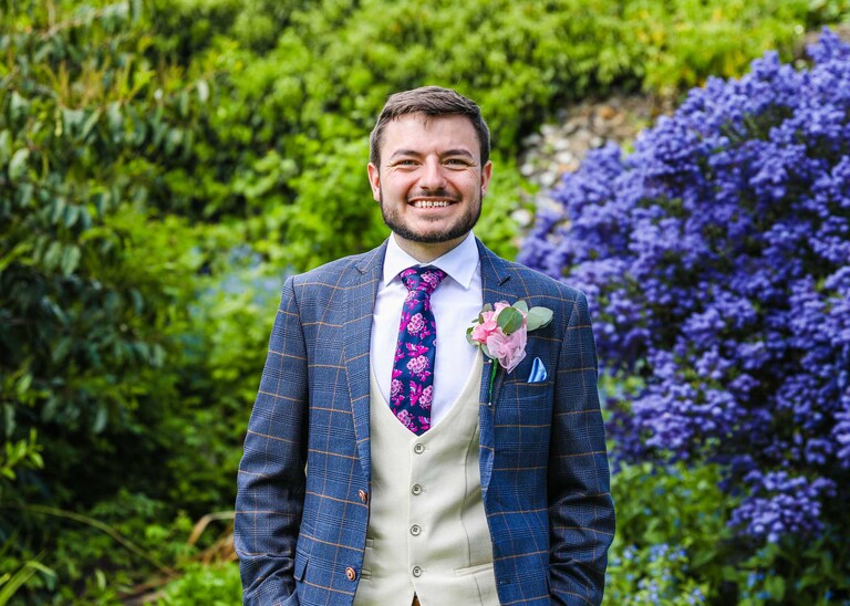 A groom smiling in front of a bright blue flowering ceanothus bush in the Abbey Gardens in Bury St Edmunds on a wedding day in Suffolk captured by Suffolk Wedding Photographers Hayley Denston Photography