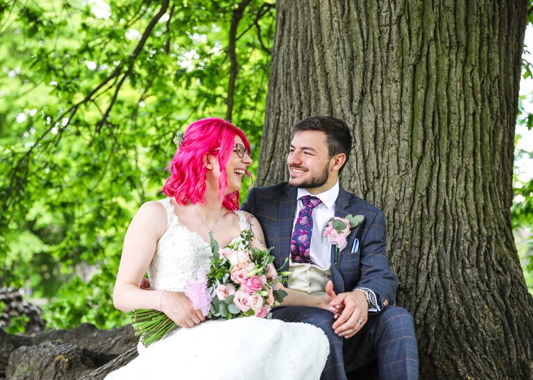 a bride and groom laughing whilst sat under an oak tree in the Abbey Gardens in Bury St Edmunds on a wedding day in Suffolk captured by Suffolk Wedding Photographers Hayley Denston Photography