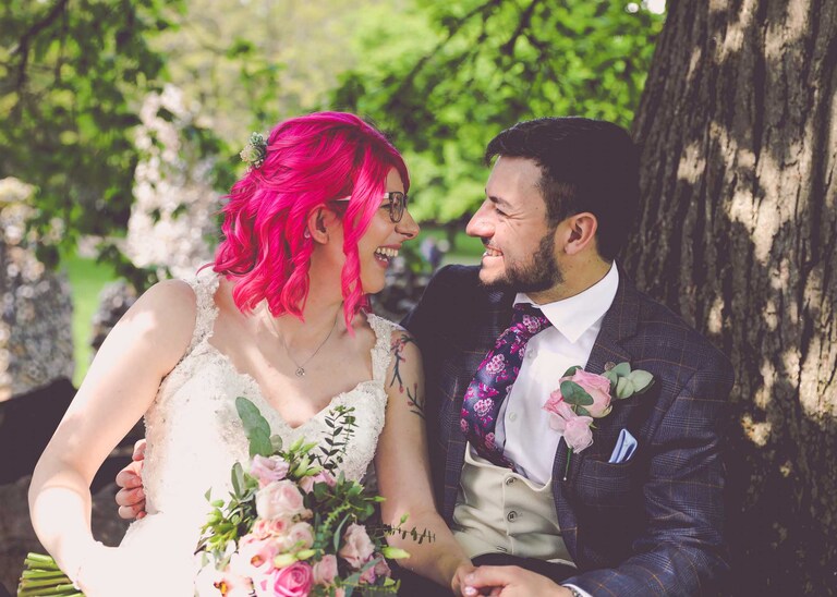 A bride and groom laughing at each other whilst sat under an oak tree in the Abbey Gardens in Bury St Edmunds on a wedding day in Suffolk captured by Suffolk Wedding Photographers Hayley Denston Photography