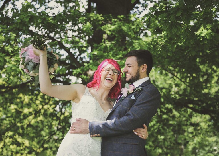 A bride and groom under a big oak tree with the bride swinging her bouquet in the Abbey Gardens in Bury St Edmunds on a wedding day in Suffolk captured by Suffolk Wedding Photographers Hayley Denston Photography