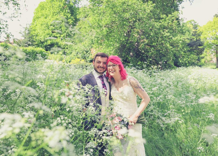 A bride and groom stood in the Abbey Gardens in Bury St Edmunds in a flowerbed full of cow parsley on a wedding day in Suffolk captured by Suffolk Wedding Photographers Hayley Denston Photography