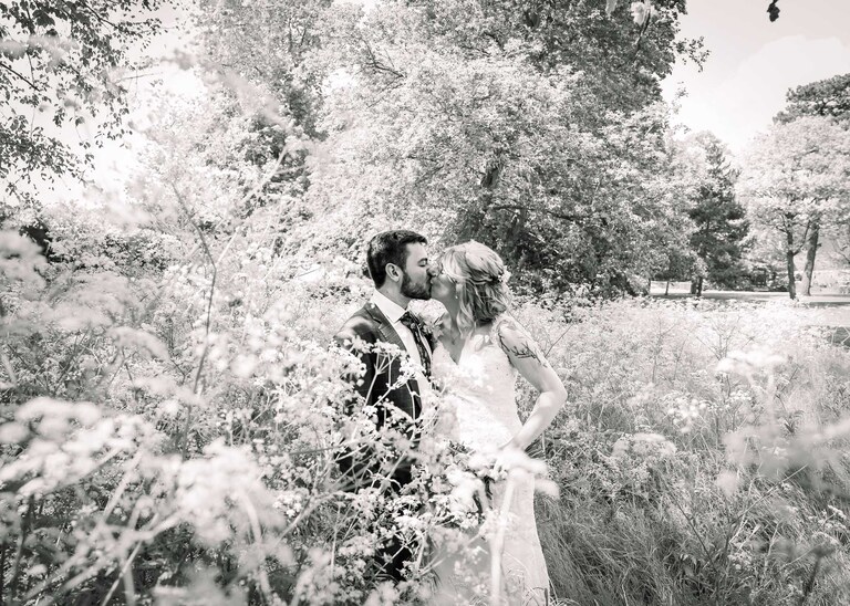 A bride and groom kissing in the Abbey Gardens in Bury St Edmunds in a flowerbed full of cow parsley on a wedding day in Suffolk captured by Suffolk Wedding Photographers Hayley Denston Photography