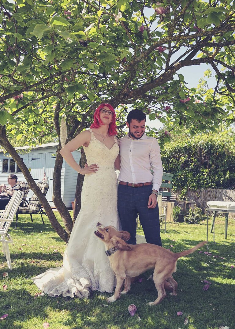 A bride and groom and a dog with zoomies underneath a magnolia tree on a wedding day in Suffolk captured by Suffolk Wedding Photographers Hayley Denston Photography