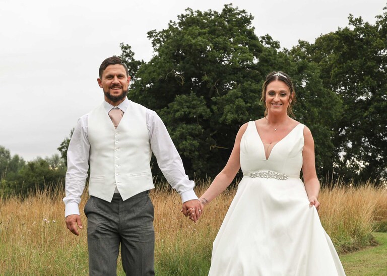 A bride and groom holding hands and smiling in the meadow with oak trees behind them at Easton Grange on a Wedding Day photographed by Suffolk Wedding Photographers Hayley Denston Photography