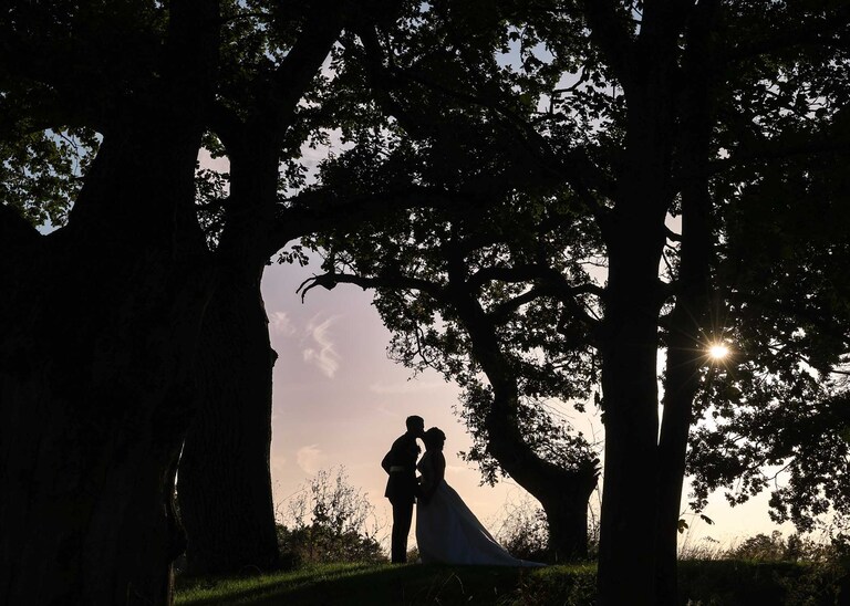 A bride and groom silhouetted and kissing at sunset under trees on their wedding day at Hungarian Hall on a Wedding Day photographed by Suffolk Wedding Photographers Hayley Denston Photography