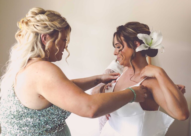 A bridesmaid helping the bride into her dress at Hungarian Hall on a Wedding Day photographed by Suffolk Wedding Photographers Hayley Denston Photography