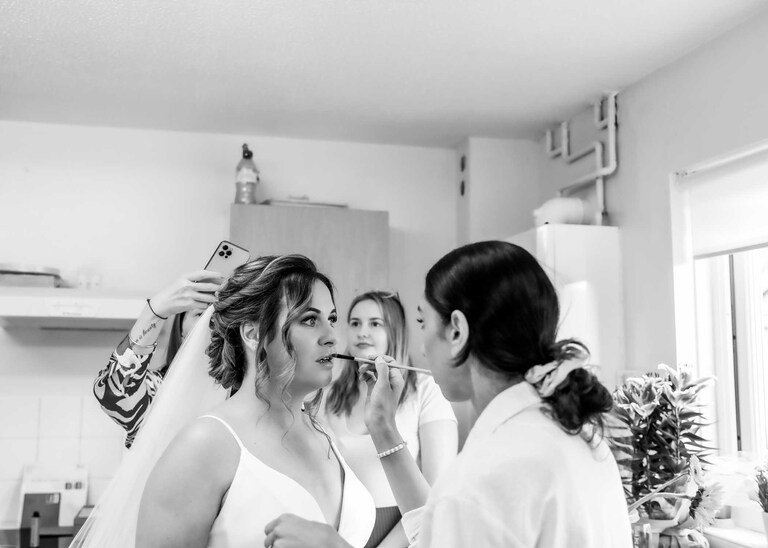 A bride having her lipstick done in the kitchen whilst the hairdresser takes a photo of her hair at Hungarian Hall on a Wedding Day photographed by Suffolk Wedding Photographers Hayley Denston Photography