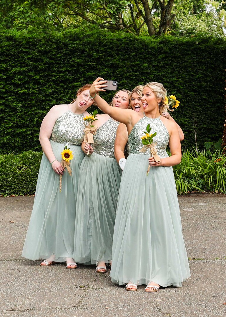 Bridesmaids dressed in sage green dresses with sparkly sequins pulling faces and taking a selfie before the wedding ceremony at Hungarian Hall on a Wedding Day photographed by Suffolk Wedding Photographers Hayley Denston Photography