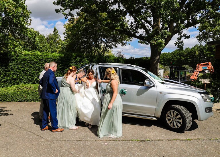 A bride getting out of a vehicle being helped by her bridesmaids and dad before her outdoor wedding ceremony at Hungarian Hall on a Wedding Day photographed by Suffolk Wedding Photographers Hayley Denston Photography
