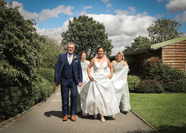 A bride and her dad walking whilst the bridesmaids carry her train towards her outdoor wedding ceremony at Hungarian Hall on a Wedding Day photographed by Suffolk Wedding Photographers Hayley Denston Photography