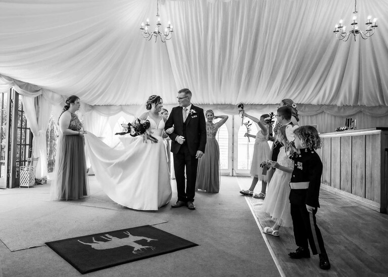 A bride smiling and looking at her dad whilst the bridal party surround them in a marquee before her wedding ceremony at Hungarian Hall on a Wedding Day photographed by Suffolk Wedding Photographers Hayley Denston Photography