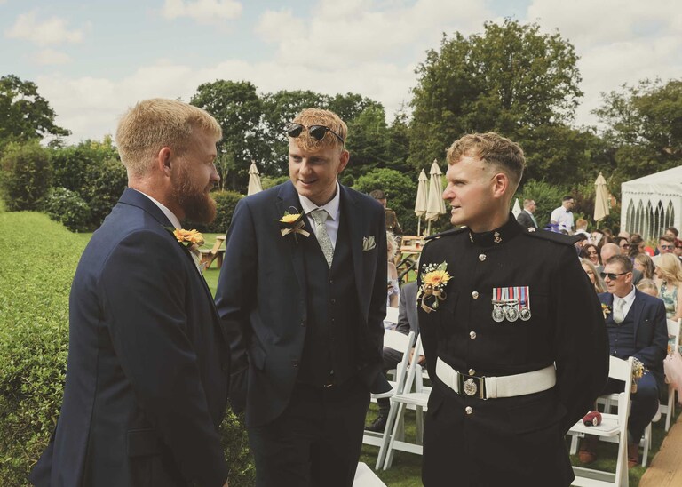 A groom chatting with his groomsmen at the top of the aisle before his wedding ceremony at Hungarian Hall on a Wedding Day photographed by Suffolk Wedding Photographers Hayley Denston Photography