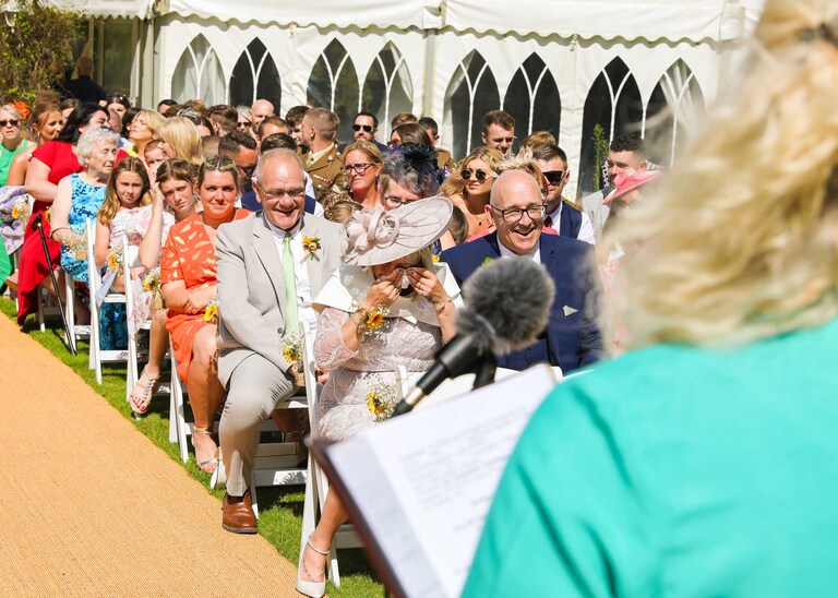 A mother of the bride wiping away her tears before her daughter walks up the aisle for an outdoor wedding ceremony at Hungarian Hall on a Wedding Day photographed by Suffolk Wedding Photographers Hayley Denston Photography