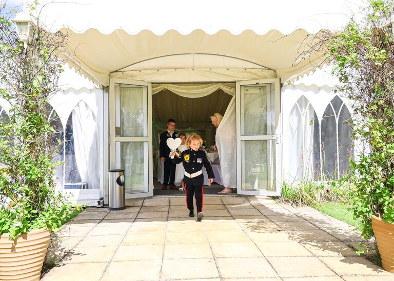 A page boy about to lead the bridal procession up the aisle during an outdoor wedding ceremony at Hungarian Hall on a Wedding Day photographed by Suffolk Wedding Photographers Hayley Denston Photography
