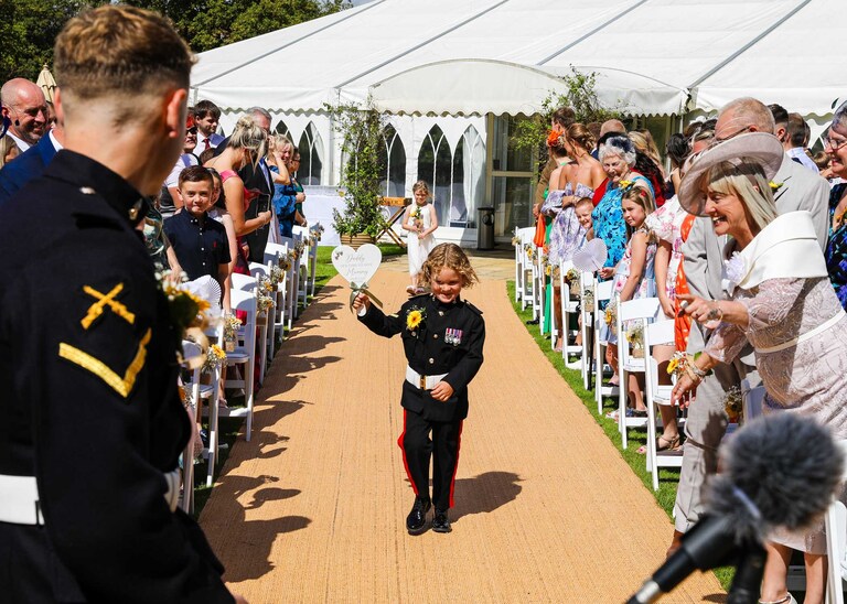 A pageboy walking up the aisle in mini ones uniform with a sign reading daddy it's time to give mummy our last name at Hungarian Hall on a Wedding Day photographed by Suffolk Wedding Photographers Hayley Denston Photography