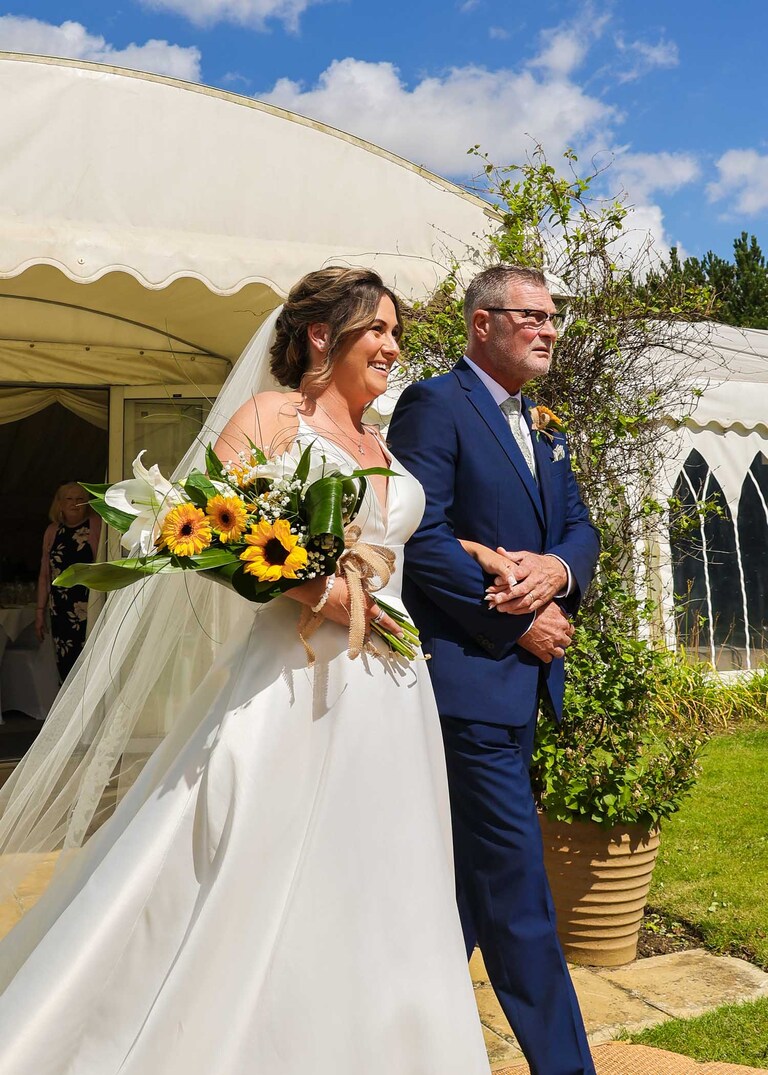 A bride and her dad about to walk up the aisle for an outdoor ceremony with blue skies behind them at Hungarian Hall on a Wedding Day photographed by Suffolk Wedding Photographers Hayley Denston Photography