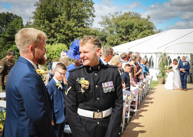 A groom smiling and winking at his bestman after seeing his bride and her dad walking up the aisle at Hungarian Hall on a Wedding Day photographed by Suffolk Wedding Photographers Hayley Denston Photography