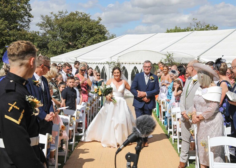 A bride and her dad walking up the aisle and the bride looking at the groom during an outdoor wedding ceremony at Hungarian Hall on a Wedding Day photographed by Suffolk Wedding Photographers Hayley Denston Photography